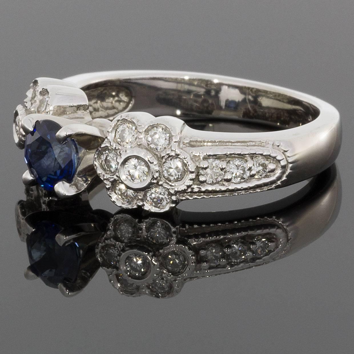 This breathtaking ring features a beautiful, .70 carat, natural, 5mm round cut, blue sapphire. This gorgeous sapphire is prong set in a 14 karat white gold diamond ring. The ring features a beautiful flower design with bezel & bead set diamonds