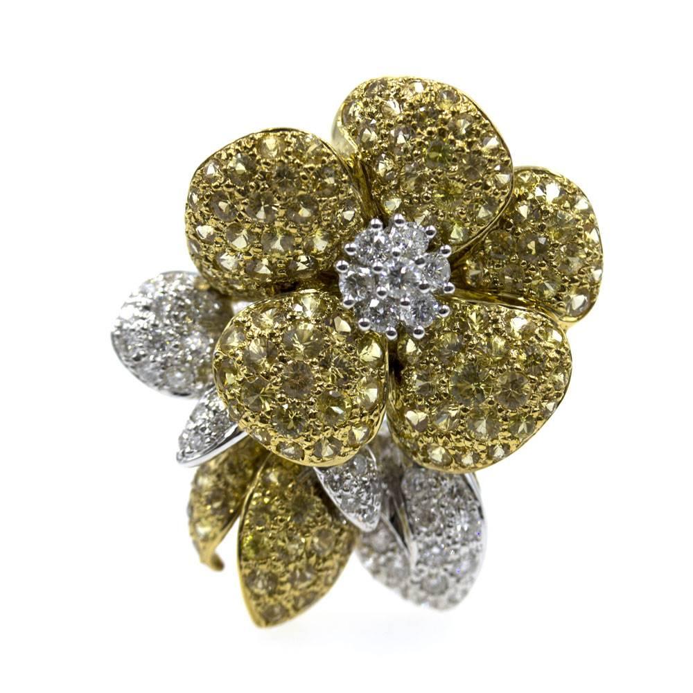 This beautiful flower pin is crafted in two-tone 18 karat yellow gold. The brooch features 1.25 carat total weight of white diamonds graded G/H color and SI clarity. Yellow sapphires create the bright petals of the flower. The pin measures 30 x