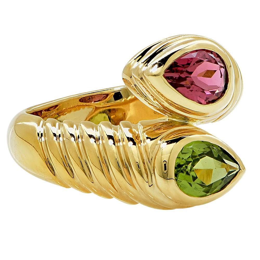 This fashionable ring by designer Bulgari features peridot and pink tourmaline gemstones. Crafted in solid 18k yellow gold, the ring has a fine polished finish with a gemstone at each end of the of the ring. One end features a pear shaped pink