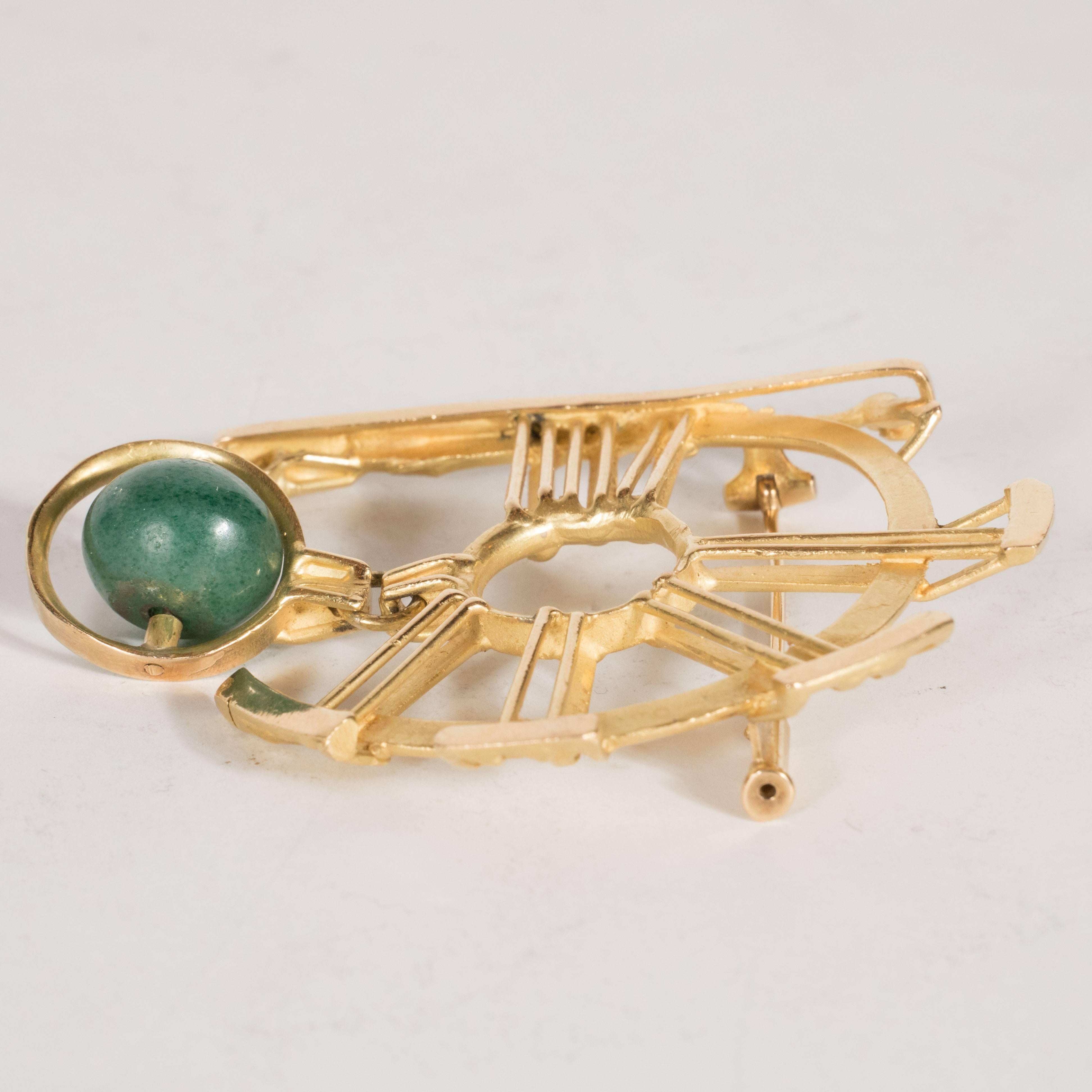 This dynamic geometric brooch features vertical stokes in 18 karat yellow gold emanating from a central circular form. A serpentine jade wheel on a horizontal axis hangs from one of these stokes, encompassed by a circle that echoes the central form.