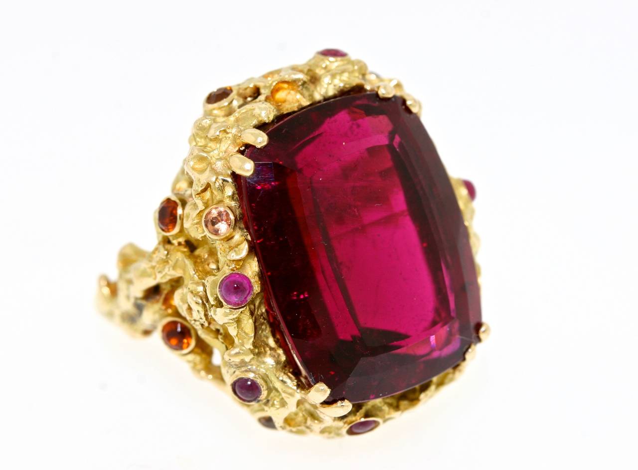 A spectacular Chaumet Paris 18k heavily textured gold ring centering a large faceted modified emerald cut rubellite stone of a deep jewel tone shade of red surrounded by sixteen faceted and cabochon stones of various complimentary shades of red,