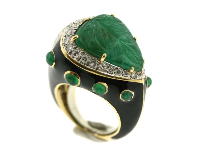 A statement David Webb ring in 18k gold centering a triangular shaped carved emerald surrounded by 40 brilliant cut diamonds and 8 cabochon emeralds in a setting of jet black enamel. Webb often used one-of-a-kind stones to create unusual and stylish