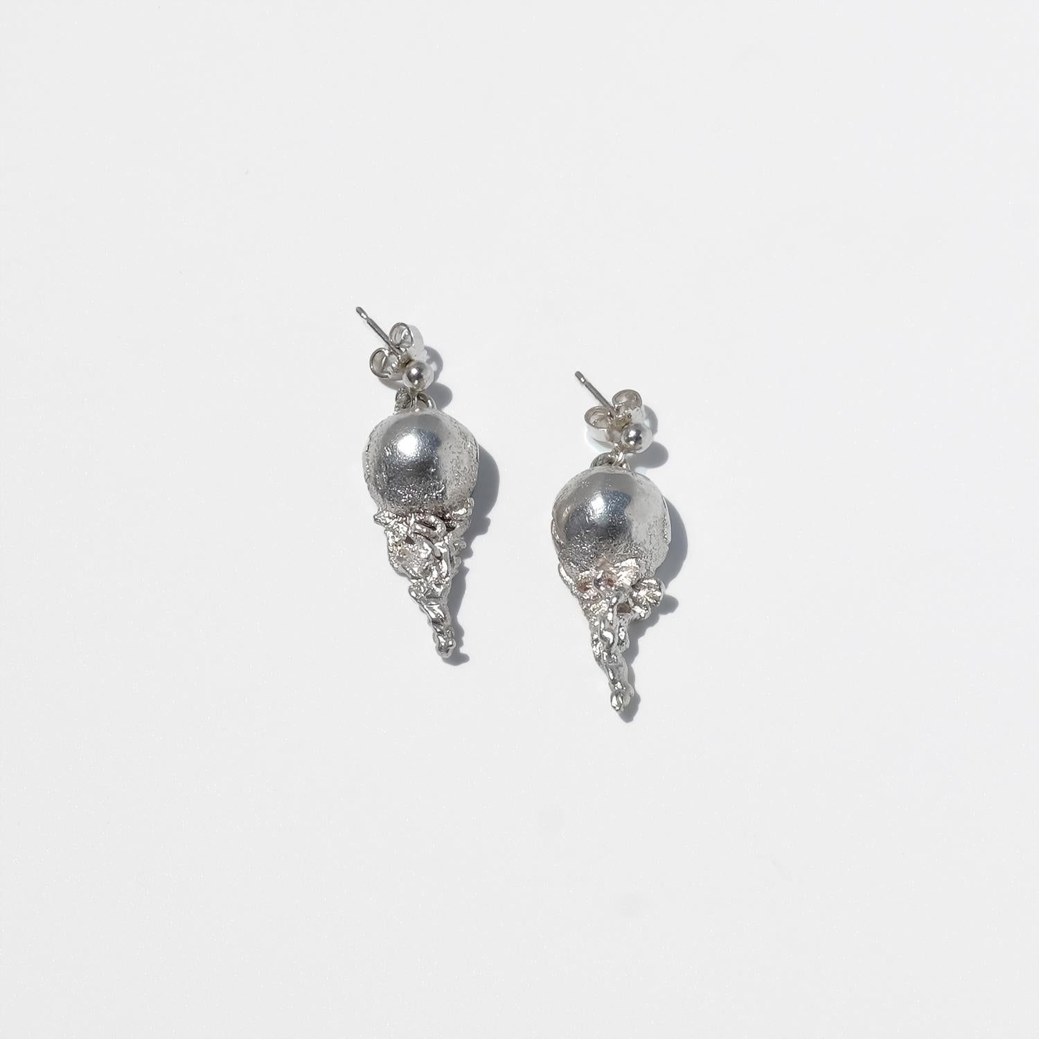 Questi orecchini a perno sono realizzati in argento e presentano una sfera d'argento da cui parte una forma organica e strutturata che si estende verso il basso. La sfera si trova vicino al lobo dell'orecchio, ancorando il design, mentre la parte