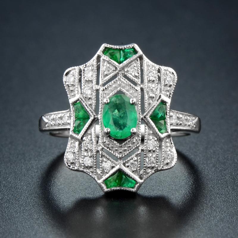 Una brillante esmeralda verde vibrante es la corona de esta soberbia y deslumbrante joya fabricada a mano, creada en estilo Art Déco clásico. La magnífica gema está acentuada con diamantes y esmeraldas de talla francesa en su parte superior,