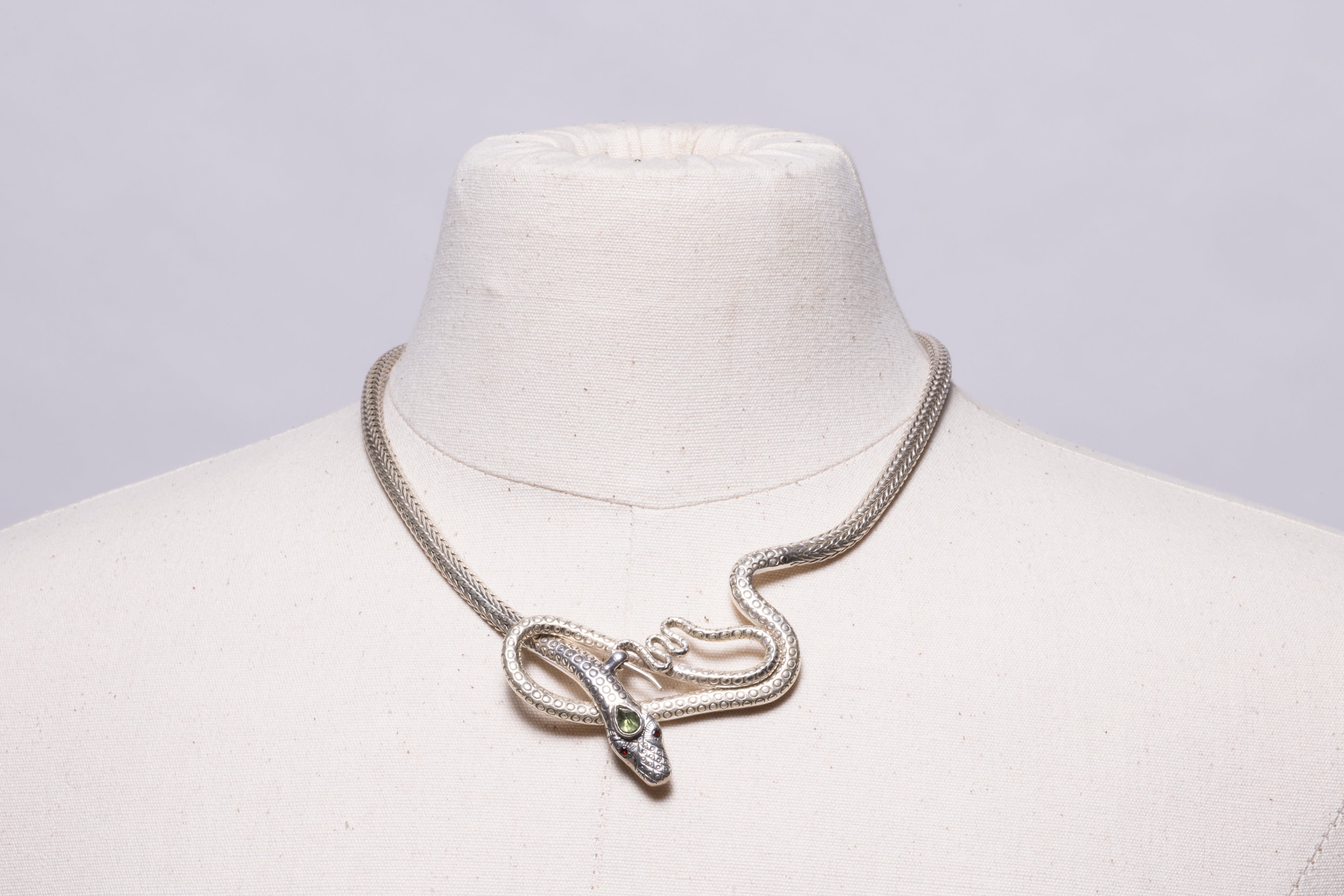A condensed herringbone pattern snake chain necklace with a looping snake at the front with faceted garnet eyes and a faceted, pear-shaped third eye in peridot.  The clasp is in the front.