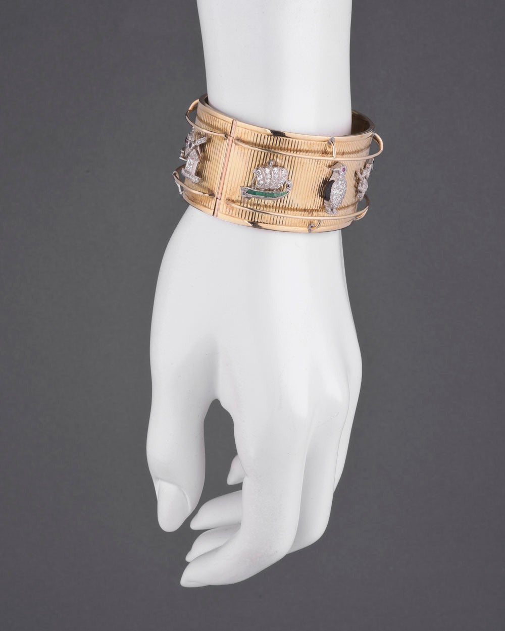 Wide oval-shaped cuff bracelet, set with eight gem-set and platinum Art Deco charms spaced evenly around the cuff, the charms including a stork carrying a baby, kangaroo, tea pot, windmill, boat, penguin, witch and bellhop, the cuff in 14k yellow