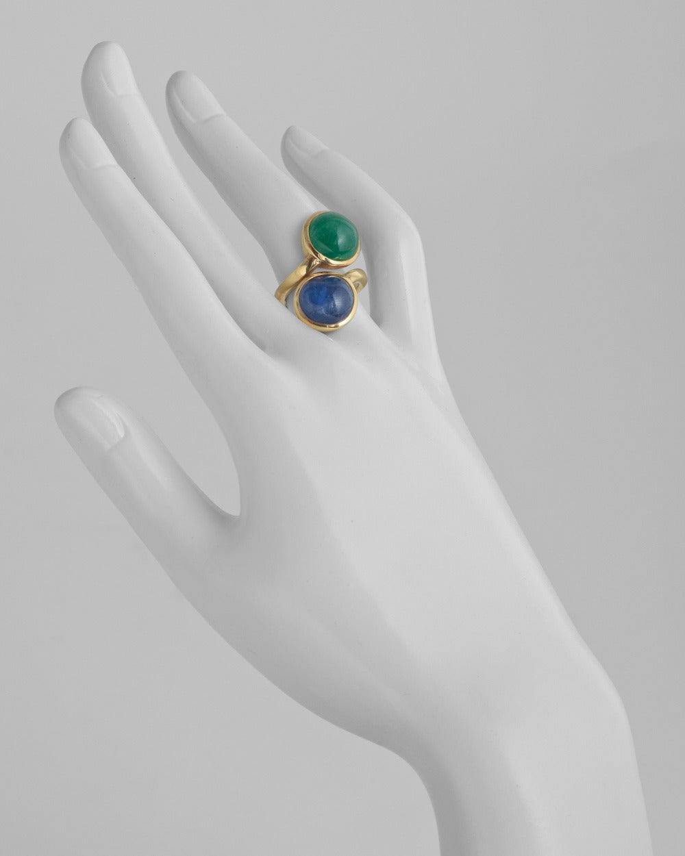 Double cabochon bypass ring, showcasing a cabochon-cut emerald and sapphire weighing approximately 7.66 carats and 8.88 carats, respectively, either gemstone measuring approximately 12mm in diameter from the top, mounted in 18k yellow gold, circa