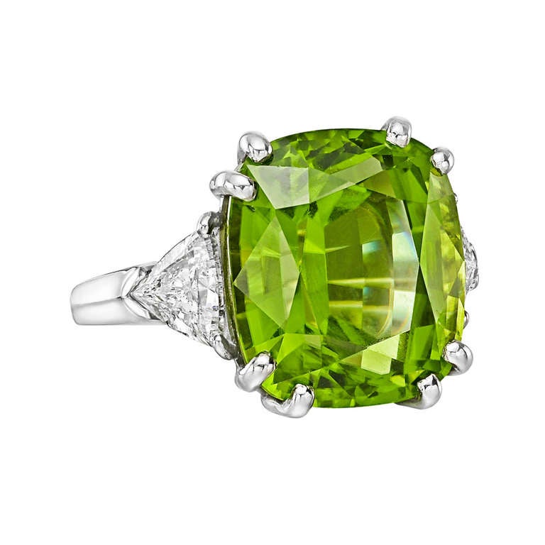 Peridot and diamond cocktail ring, centering on a cushion-cut peridot weighing approximately 18.98 carats, flanked by two trillion-cut diamond shoulders weighing approximately 1.44 total carats, mounted in platinum. Re-sizable.