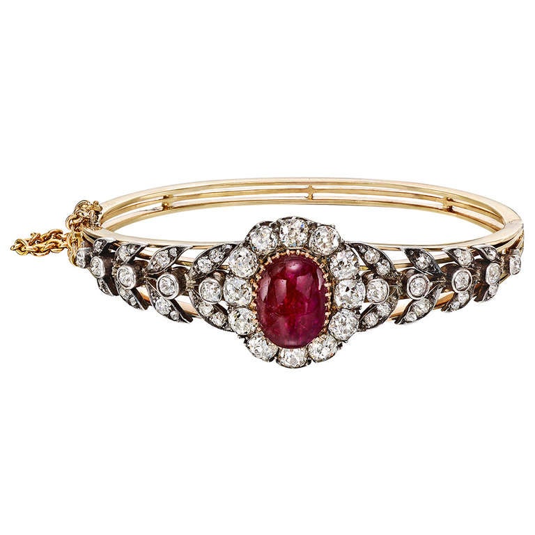 Antique gem-set oval bangle bracelet, centering on an oval-shaped cabochon ruby surrounded by twelve larger old mine cut diamonds weighing approximately 3.00 total carats and flanked by diamond-set foliate-style accents, the smaller diamonds