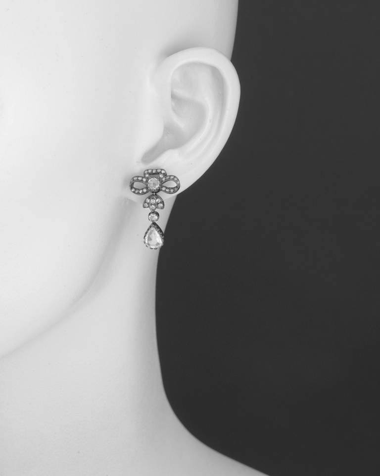 Antique-style diamond bow short drop earrings, designed with a pavé diamond bow surmount centering a circular-shaped rose-cut diamond, suspending a smaller circular-shaped rose-cut diamond to a larger pear-shaped rose-cut diamond, the diamonds