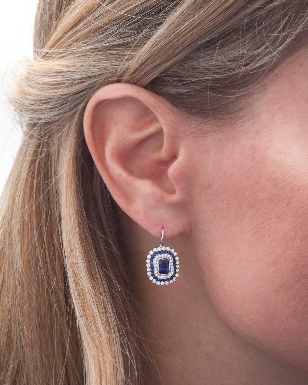 Sapphire and diamond cluster drop earrings, centering a larger emerald-cut sapphire surrounded by round-cut diamonds to a row of calibre-cut sapphires and round-cut diamond scalloped edge, mounted in platinum. Two emerald-cut sapphires weighing 1.34