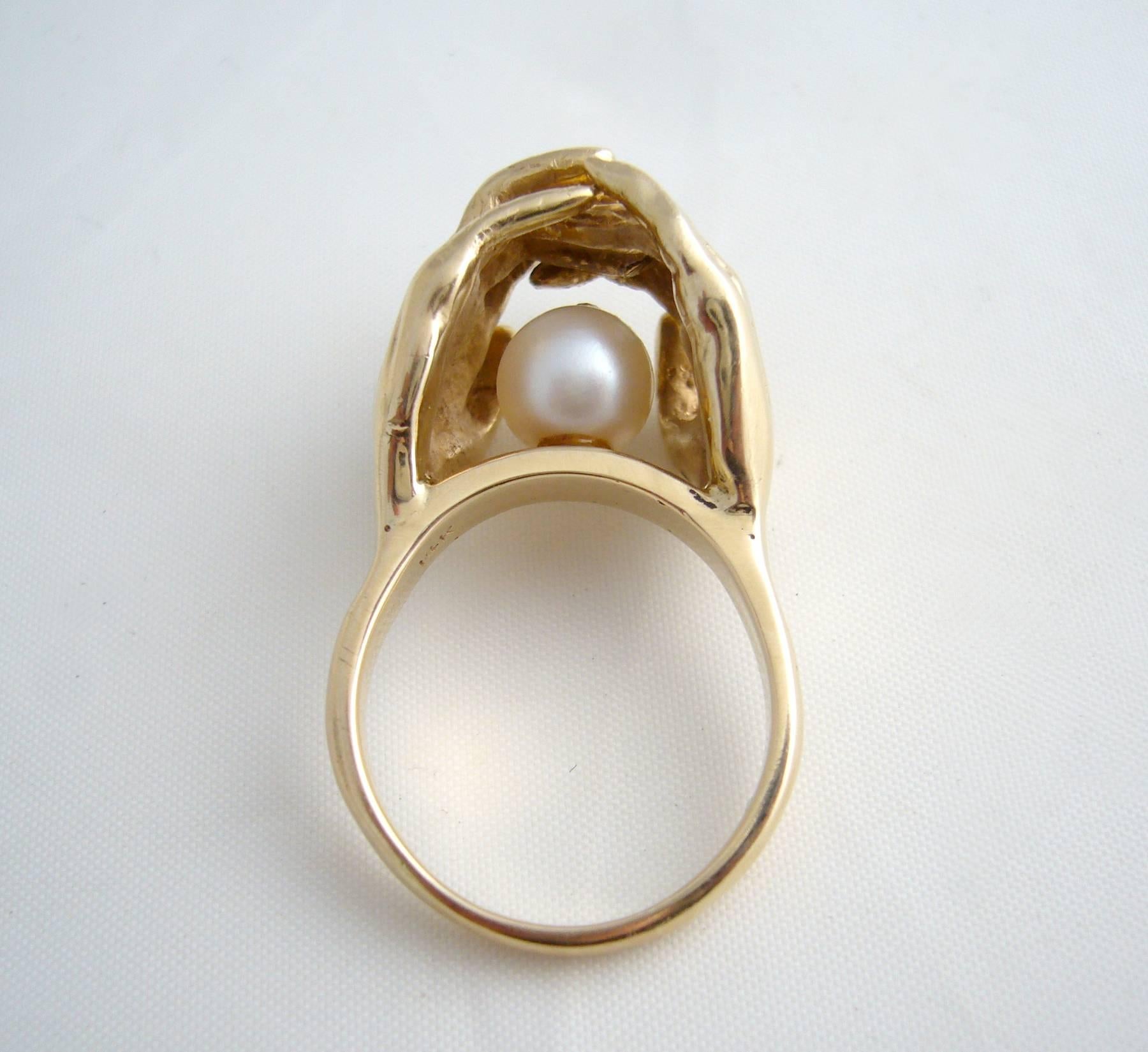 Gold and pearl surrealist ring depicting two very detailed hands holding a pearl, signifying love or deep friendship.  Ring is a finger size 7.5 and weighs 14.6 grams.  A cool, unconventional alternate to a modern day engagement or wedding ring.