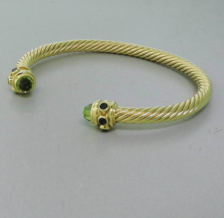Classic David Yurman cable cuff bracelet in 18K yellow gold featuring peridot and sapphires. Cable measures 5mm in diameter, ends are 9.2mm at widest points. Cuff is slightly flexible and will fit up to 7 1/2