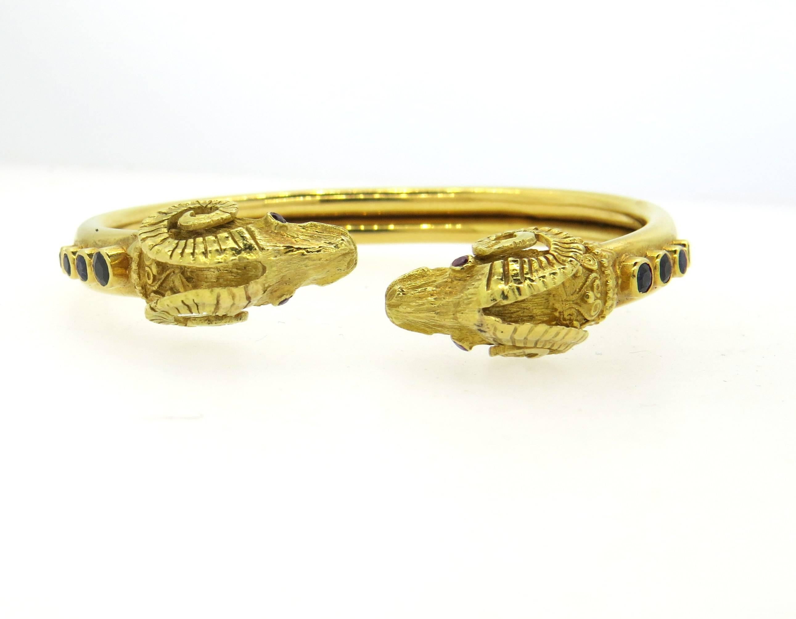 AN 18k yellow gold bangle, crafted by Ilias Lalaounis, featuring two ram's heads, decorated with blue sapphires and ruby eyes. Bracelet will fit up to 7