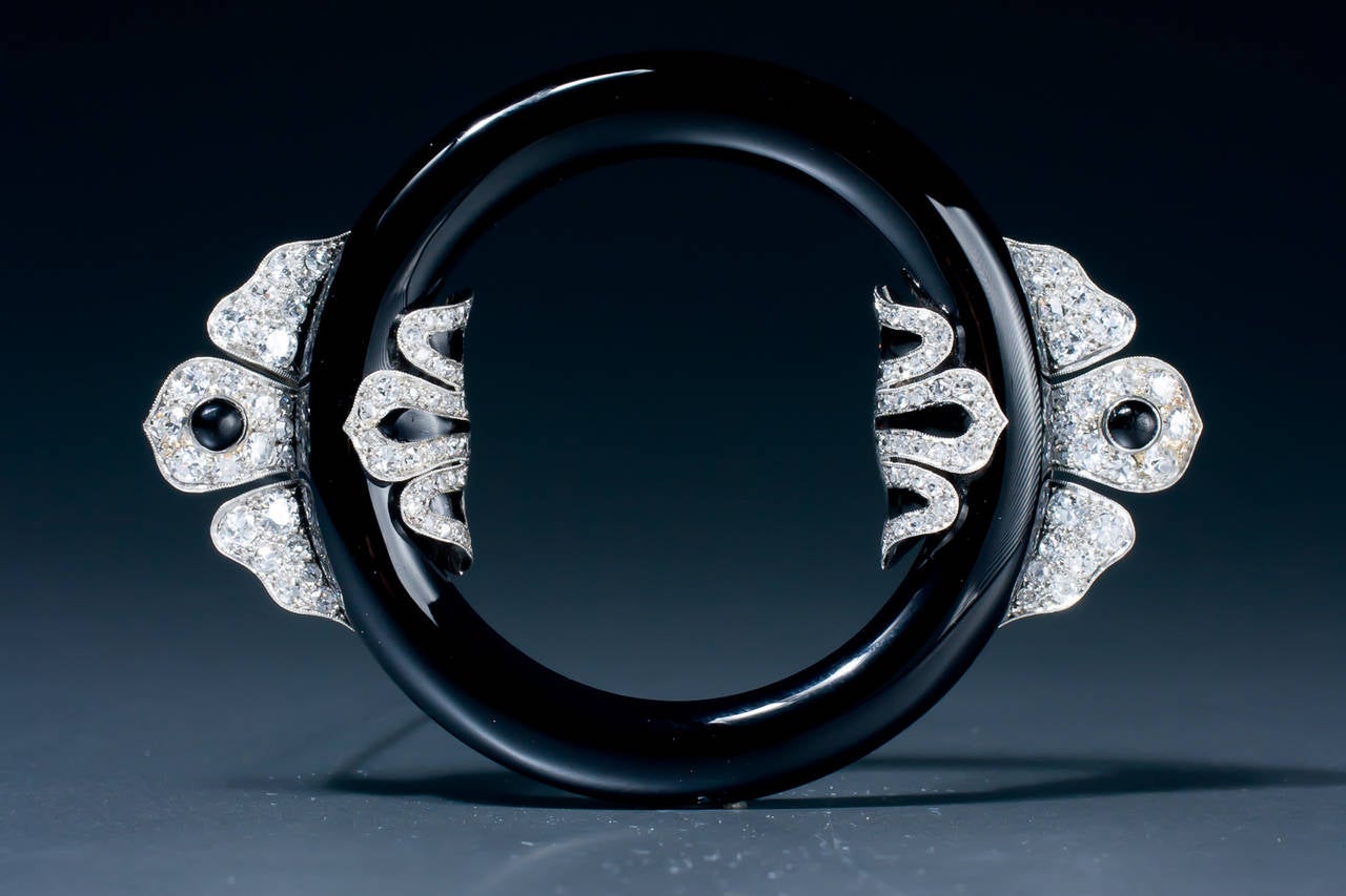 A unique early Art Deco onyx, black enamel and diamond circle brooch, designed as an onyx circle, flanked by two diamond stylized leaf shaped motifs, decorated with black enamel and onyx cabochon, mounted in with 