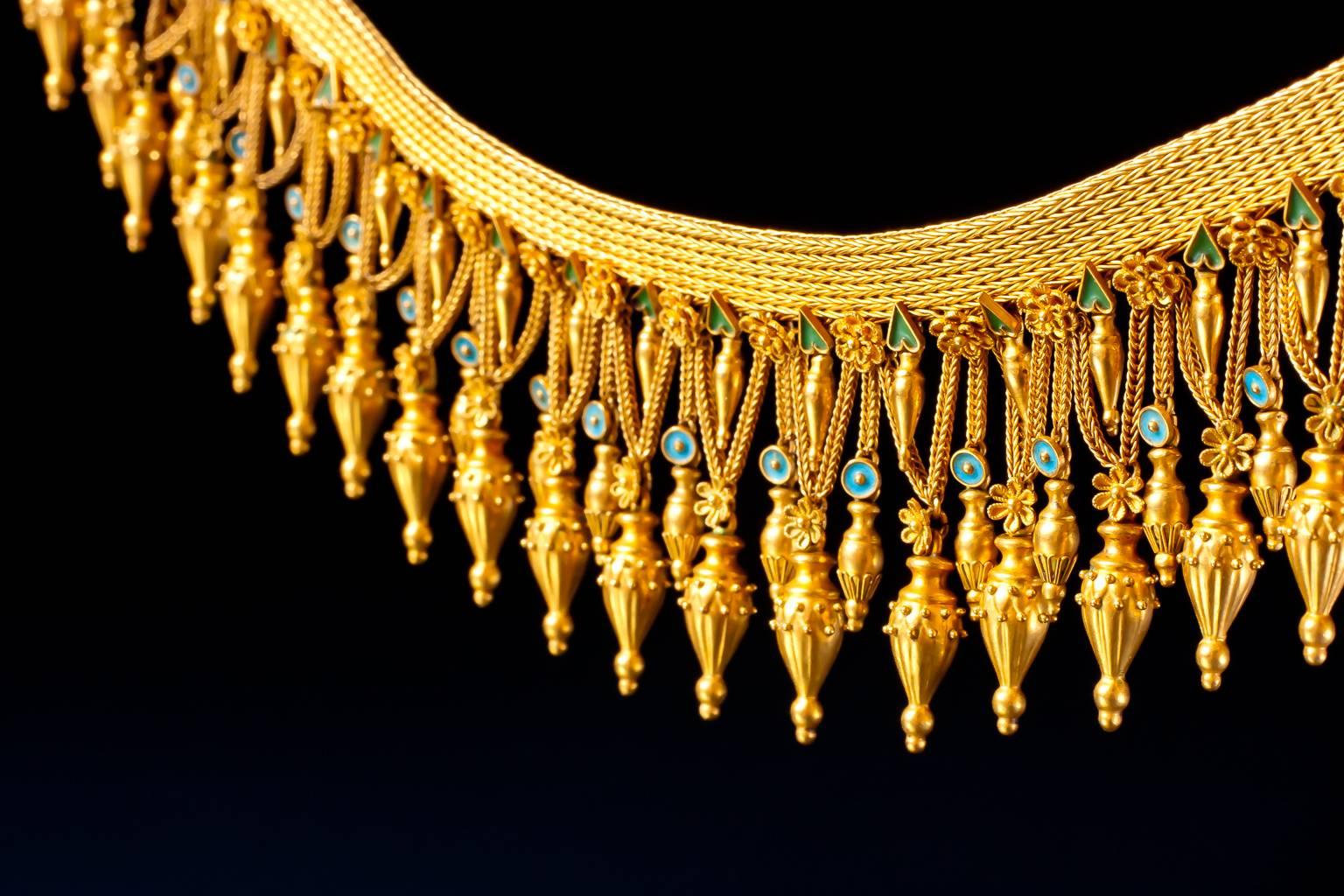An important revivalist gold and enamel 'Melos' necklace,
the flexible band of woven gold suspending a festooned fringe of stylised amphora drops alternately plain or beautifully decorated with filigree and granulation.The chain suspensions further