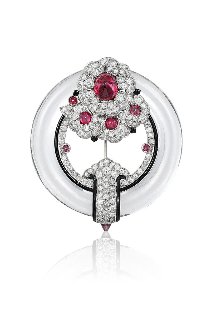 A beautifully crafted pin depicting a cluster of four flowers centered around fine cabochon rubies placed on a hand carved round tube of rock crystal, all pavé set with diamonds and with enamel detailing.

Possibly a Cartier piece due to its style,