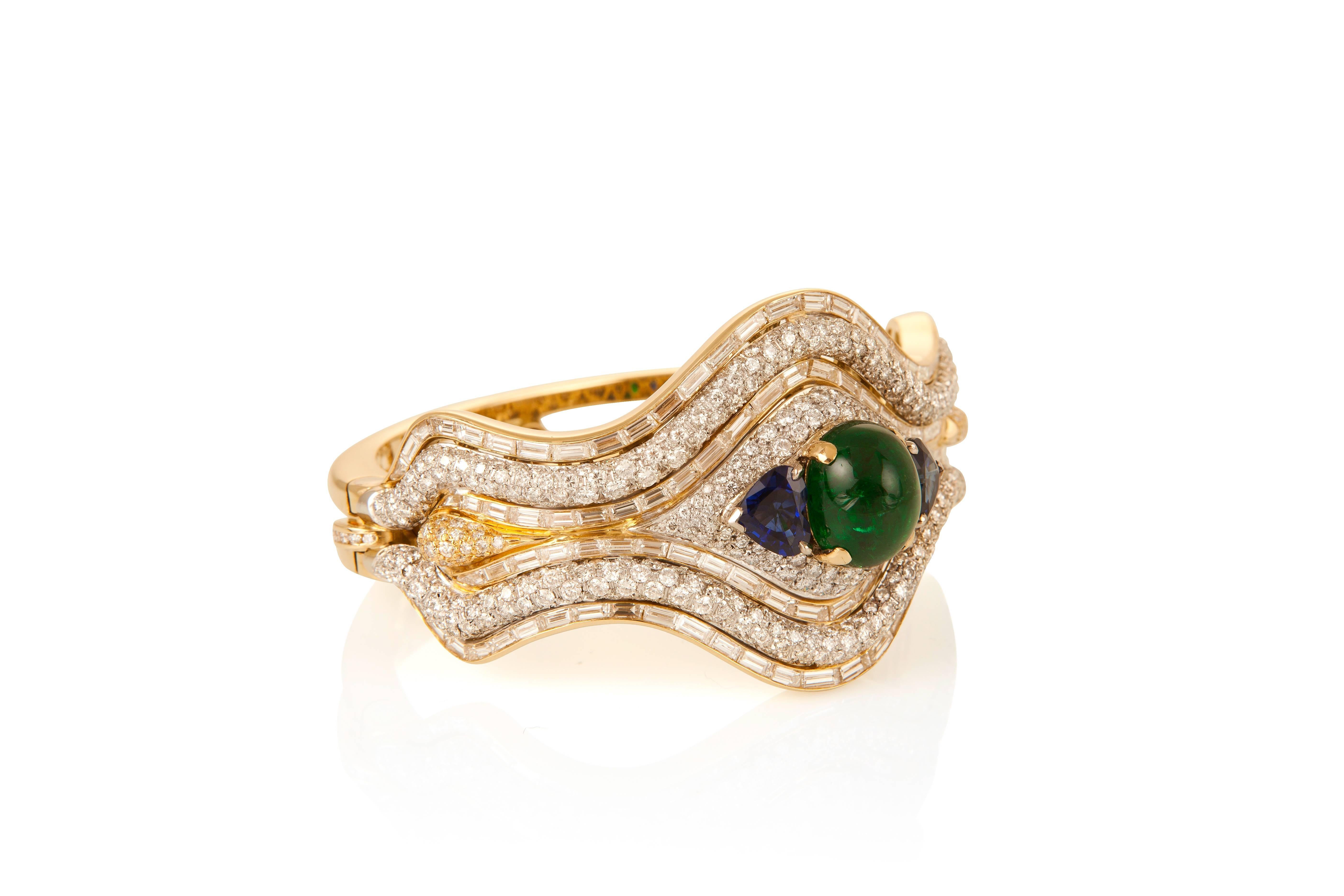 This grandly proportioned bangle features an emerald cabochon weighing 10.70 carats, two heart shape sapphires weighing 4.79 carats, and round and baguette diamonds in a flowing design, all mounted in 18 karat yellow gold.