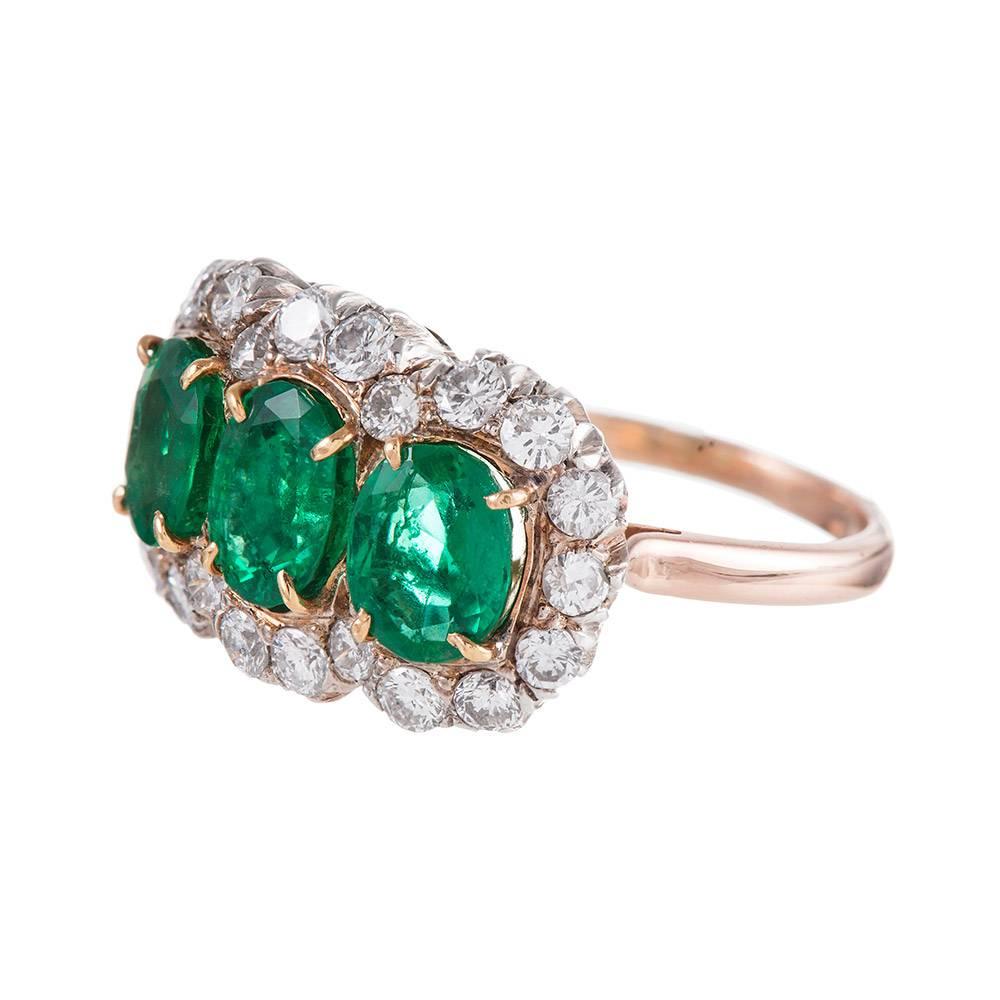 14k white and yellow gold ring set with three oval emeralds and framed by a cluster of white diamonds. Each emerald weighs approximately 1 carat and the twenty-two diamonds weigh 1.00 carat combined. This ring has an impressive presence on the