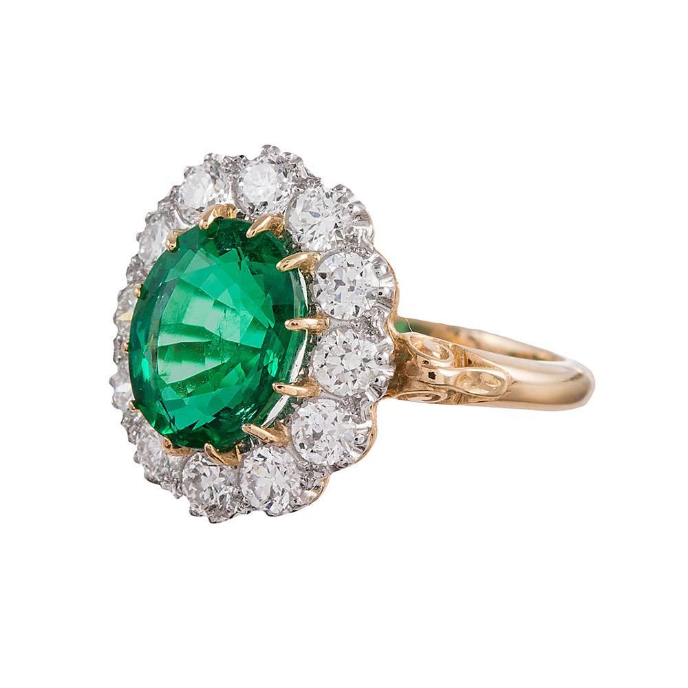 A classic “Princess Diana” style cluster ring, rendered in platinum and 18 karat yellow gold. The center is set with a 2.73 carat oval brilliant emerald and framed by 1.16 carats of white diamonds. This ring is of a classic vintage design, but of