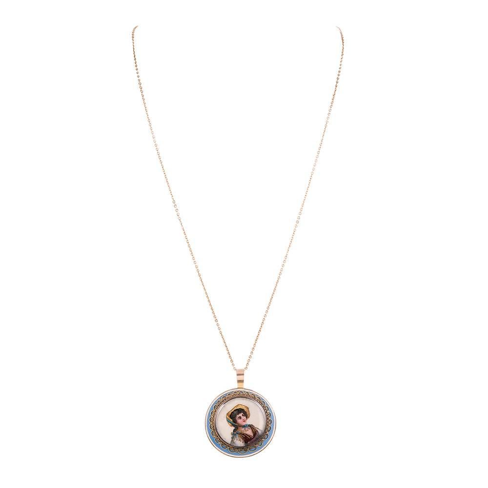 A 1.25 inch diameter ornament with periwinkle blue and white enamel framing a miniature portrait of a woman. Her image is further bordered by a golden scroll design. The back has a transparent hair locket. 