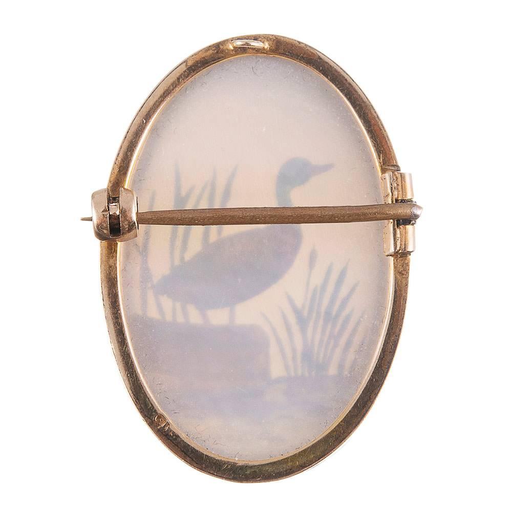 Framed in 9 carat yellow gold, the reverse painted duck stands on the shore overlooking the water, his lustrous feathers bursting with color. Water laps at his webbed feet and reeds provide the backdrop. 1 ¼ by 7/8 inches oval and painted on mother