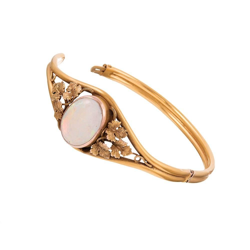 14k yellow gold oval shaped bangle bracelet, opening with a single hinge and finished with a safety chain. The top is decorated with a large opal cabochon and flanked on each side by a trio of golden grape leaves. 2.5 by 2 inches internal diameter.