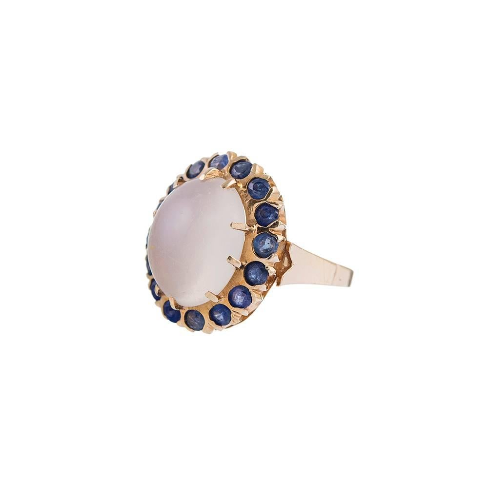 The allure of the cabochon moonstone is augmented by a frame of blue sapphires, creating a unique and wearable ornament. Rendered in 14k yellow gold, with subtle details including a stylized shank and elongated prongs, the colors lend themselves to