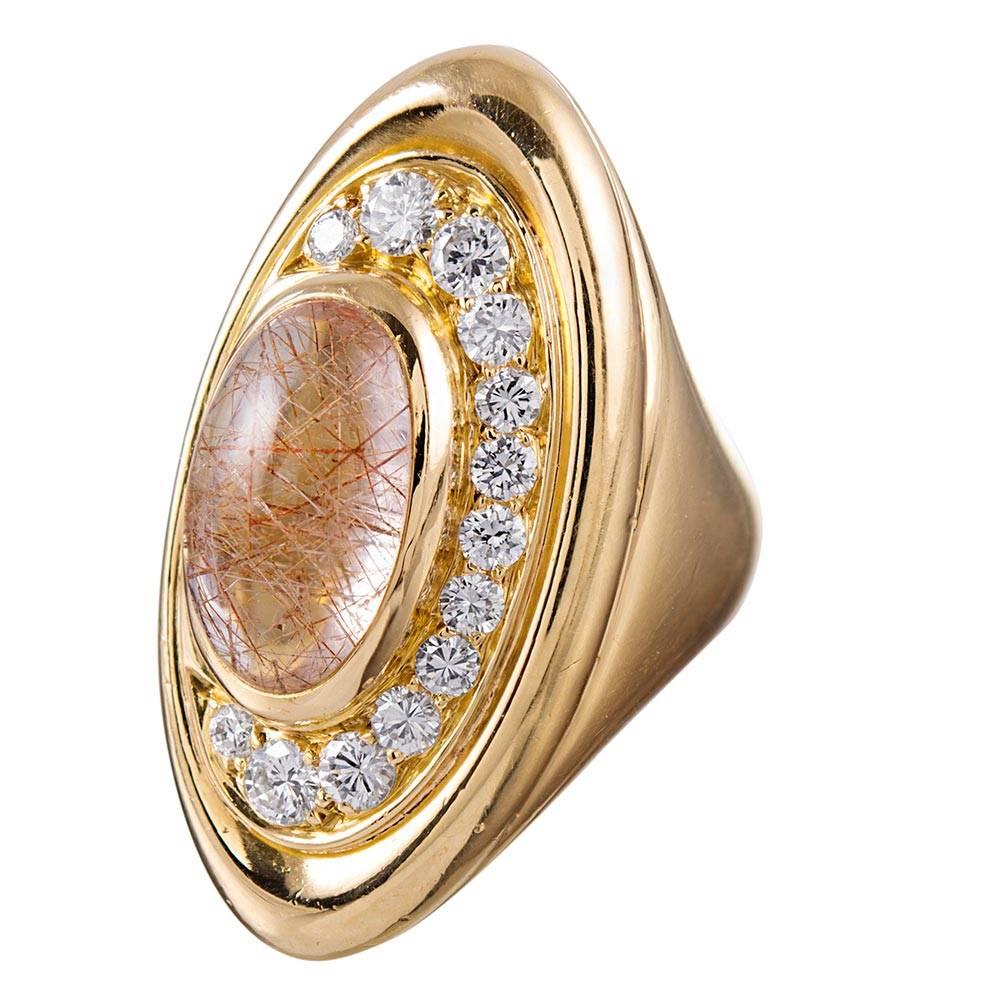Striking and ornate, the ring is designed with sweeping strokes of polished gold encircling a crescent moon shaped decoration of graduated diamonds and a cabochon rutilated quartz. The brilliant white diamonds weigh .89 carats while the center
