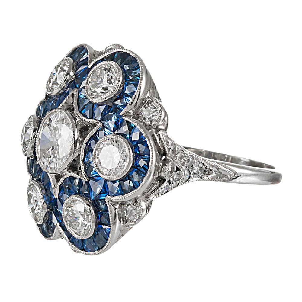 An elaborate rendering of art deco splendor, the ring is hand made in platinum and set with 2.52 carats of brilliant white diamonds, complimented by tile-set blue sapphires. Though highly authentic in appearance, this ring is of newer manufacture.