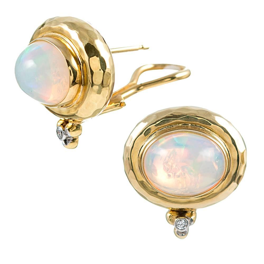 Hammered bezels of 18 karat yellow gold contain a pair of opal cabochons and are anchored at the bottom with a cluster of golden orbs and a solitary brilliant diamond. The opals boast a display of blue, green, red and purple flashes that dance in
