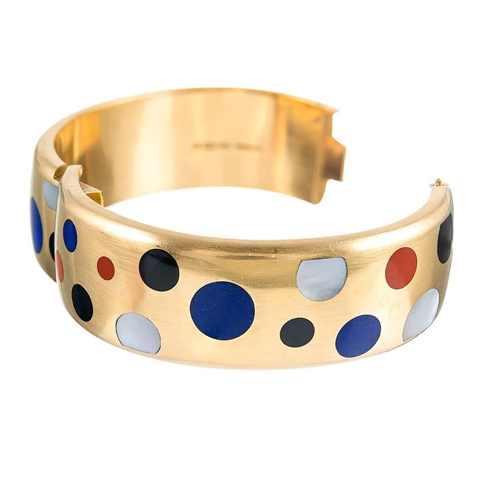 The bangle measures 7/8 of an inch wide and is rendered in 18 karat yellow gold. Decorated with a polka-dotted pattern of inlaid carnelian, lapis, onyx & mother-of-pearl, the piece offers a stylish and sophisticated look that is wearable for all