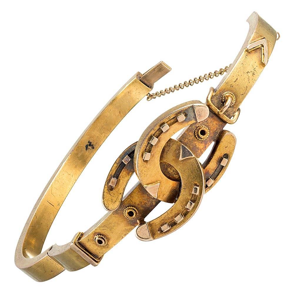 Made of 15 karat yellow gold and boasting a lovely duo of classic Victorian motifs, the bracelet is decorated with a pair of ornate horseshoes crossed atop a golden buckle. The interior diameter is 2 3/8 by 2 inches. Wear this piece alone as your