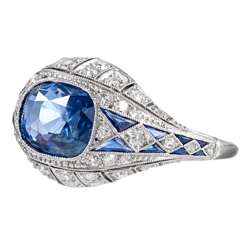 An alluring cornflower blue cushion sapphire sits nestled in an 18 karat white gold bezel, surrounded by an art deco inspired masterpiece of sapphires and brilliant diamonds. The major sapphire is accompanied by an AGL certificate stating the stone