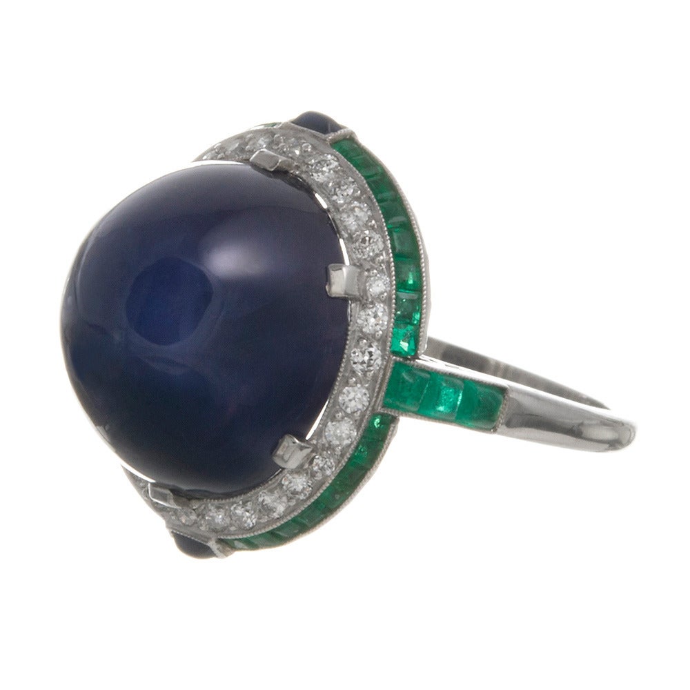 This ring has layers of interest, even beyond its stunning art deco
design. Hand made of platinum, with a subtle double frame of diamonds and
emeralds, cabochon sapphire accents peppered at each compass point. The
center stone is the ³star