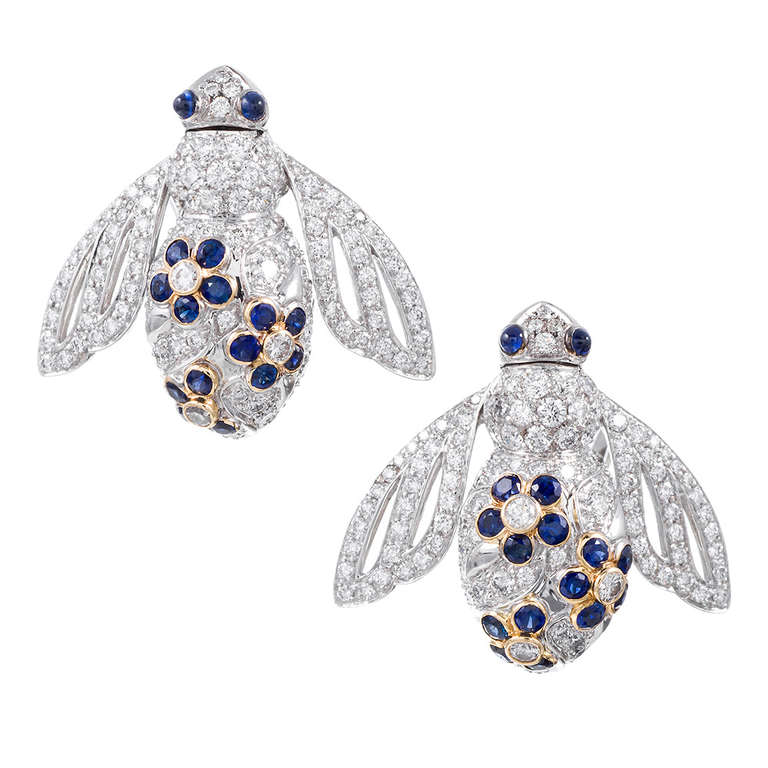 18k white gold earrings, comprised of diamonds and sapphires and shaped into the three-dimensional form of a bee. The bodies are set with 4.12 carats of brilliant white diamonds, with sapphire-studded flower petals decorating their backs. Cabochon