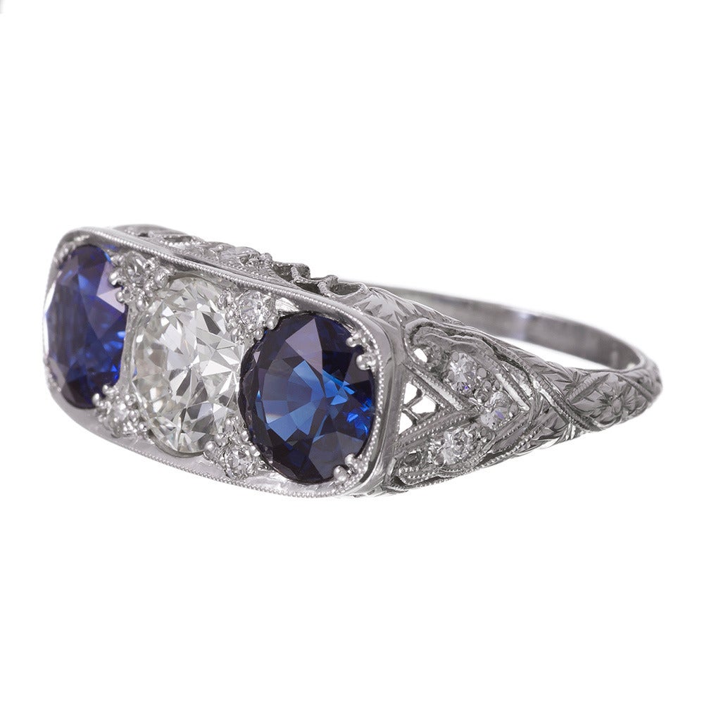 Styled after the classic Victorian English carved ring, but made during the art deco era, this beautifully-preserved three stone ring features a 1.36 carat brilliant round diamond center flanked by an ideally-matched pair of round sapphires that