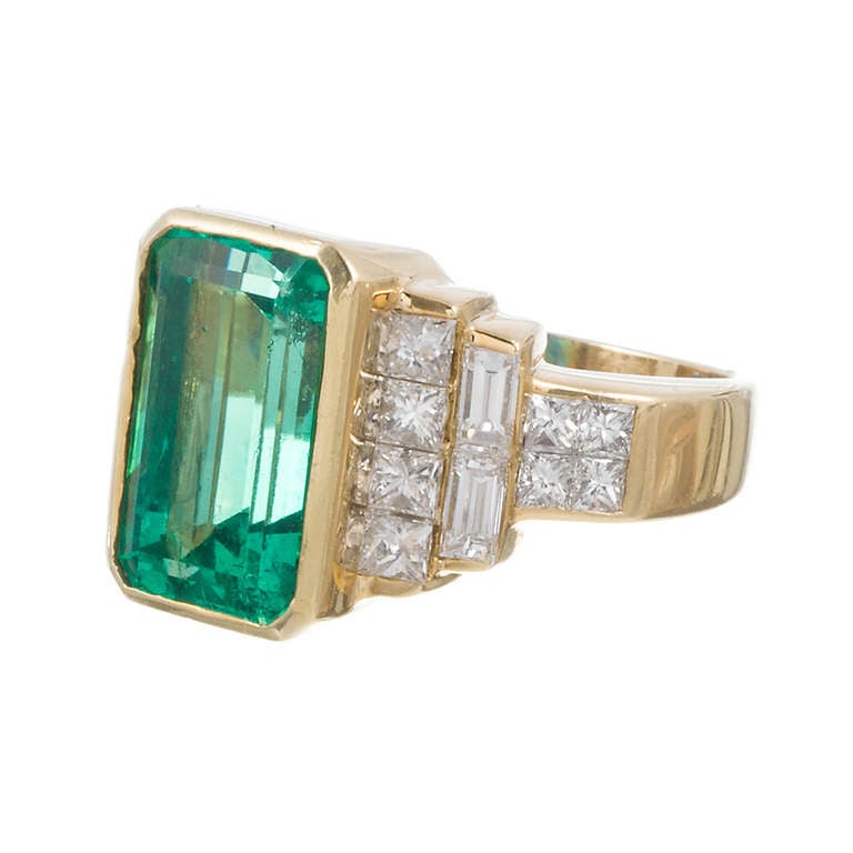 Set in 18k yellow gold and complimented by 2 carats of square- and baguette cut diamonds stacked on either side and tapering down the shank. This 7 carat emerald exhibits lovely color and is well-protected in its bezel setting.