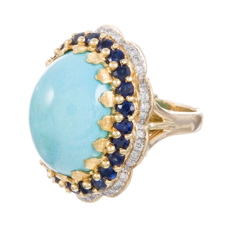 18k yellow gold ring with a substantial cabochon turquoise centerpiece, contained in unique prongs with a florentine finish and framed in a row of navy blue sapphires, then a diamond-studded scalloped border. This would make a fabulous cocktail