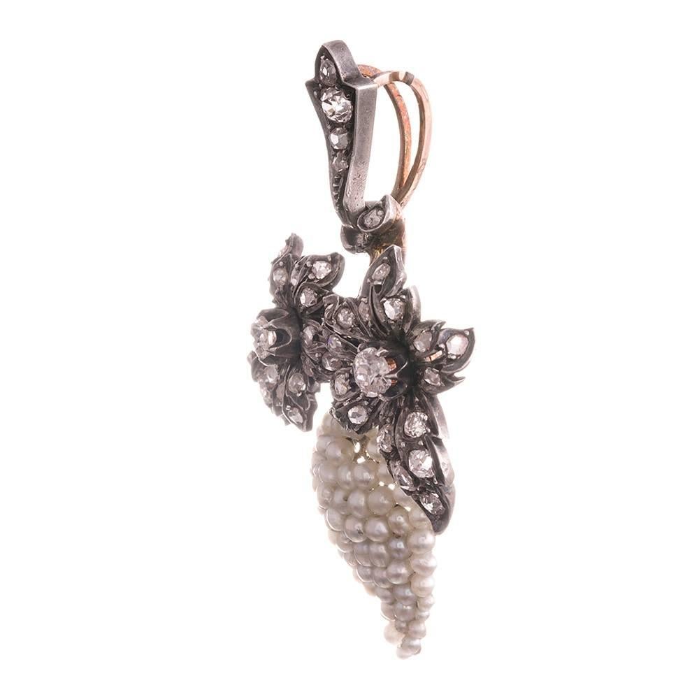 A cluster of seed pearls mimic in three dimensions the look of a bunch of grapes, set in silver over 18 karat gold. They are suspended from a pair of diamond-studded flowers and fitted with a diamond bale. In total, there are approximately 1 carat