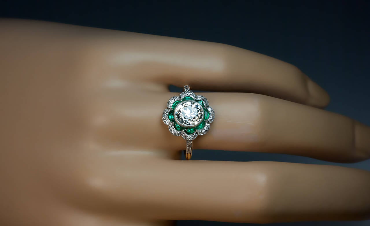 A platinum topped 18K gold ring centered with a bezel set old cut oval diamond (7 x 6.35 x 4.5 mm, approximately 1.50 ct) surrounded by eight petal shaped emeralds outlined with a curvaceous border of old cut diamonds.

Marked with French platinum