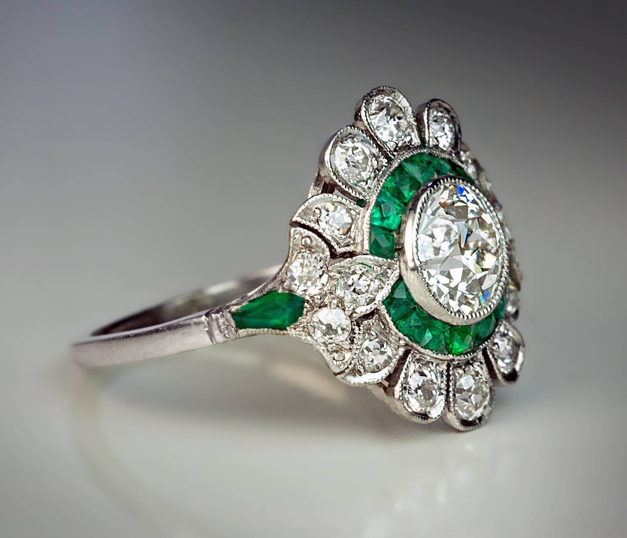 18K white gold designed as a stylized flower head flanked by fleur-de-lis shoulders, centered with an old European cut diamond (6.1-3.85 mm, approximately 1 carat) surrounded by calibre cut emeralds, estimated total diamond weight 1.34 ct

Marked