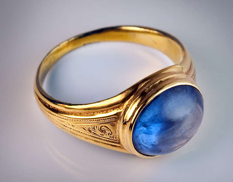 A 14K yellow gold ring with engraved decorations is bezel-set with an oval cabochon cut cornflower blue sapphire

11.5 x 9.8 x 5.8 mm, approximately 7.06 carats.

 US ring size 8  (18 mm)

The ring is marked with maker's initials ' B A ',  56