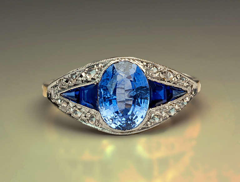 A platinum topped 18K gold ring is centered with a natural oval sapphire (7.7 x 5.9 x 4 mm, approximately 1.45 ct)  flanked by four calibre cut synthetic sapphires within a platinum frame set with rose cut diamonds.

The ring is marked with