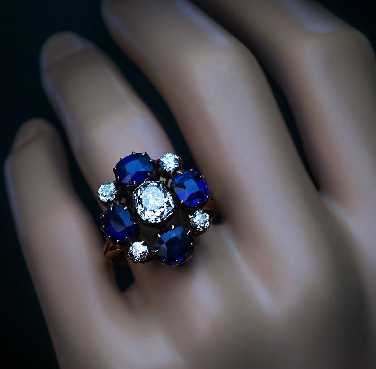 Made between 1908 and 1917.
The 14K gold ring is designed as a stylized flowerhead (20 x 19 mm), centered with an antique cushion cut diamond (7 x 6.2 x 4.8 mm, approximately 1.76 ct, I color, SI1 clarity) surrounded by four cushion cut deep blue