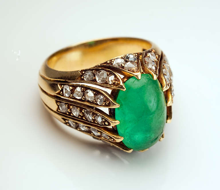 An 18K gold ribbed ring is centered with a cabochon cut emerald (13.2 x 8.9 x 4.9 mm, approximately 4.31 ct) set in a fancy bezel encrusted with 33 rose cut diamonds.

Marked with maker's initials and French eagle mark for 18K gold.

US ring