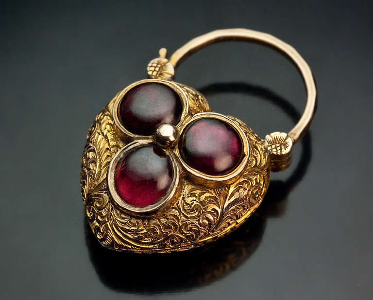 The gold puffed heart charm is finely engraved with scrolling foliage and embellished with three cabochon cut almandine garnets forming a trefoil. 

 The back of the pendant is set with a glazed miniature compartment.

 Total height 35 mm (1 3/8