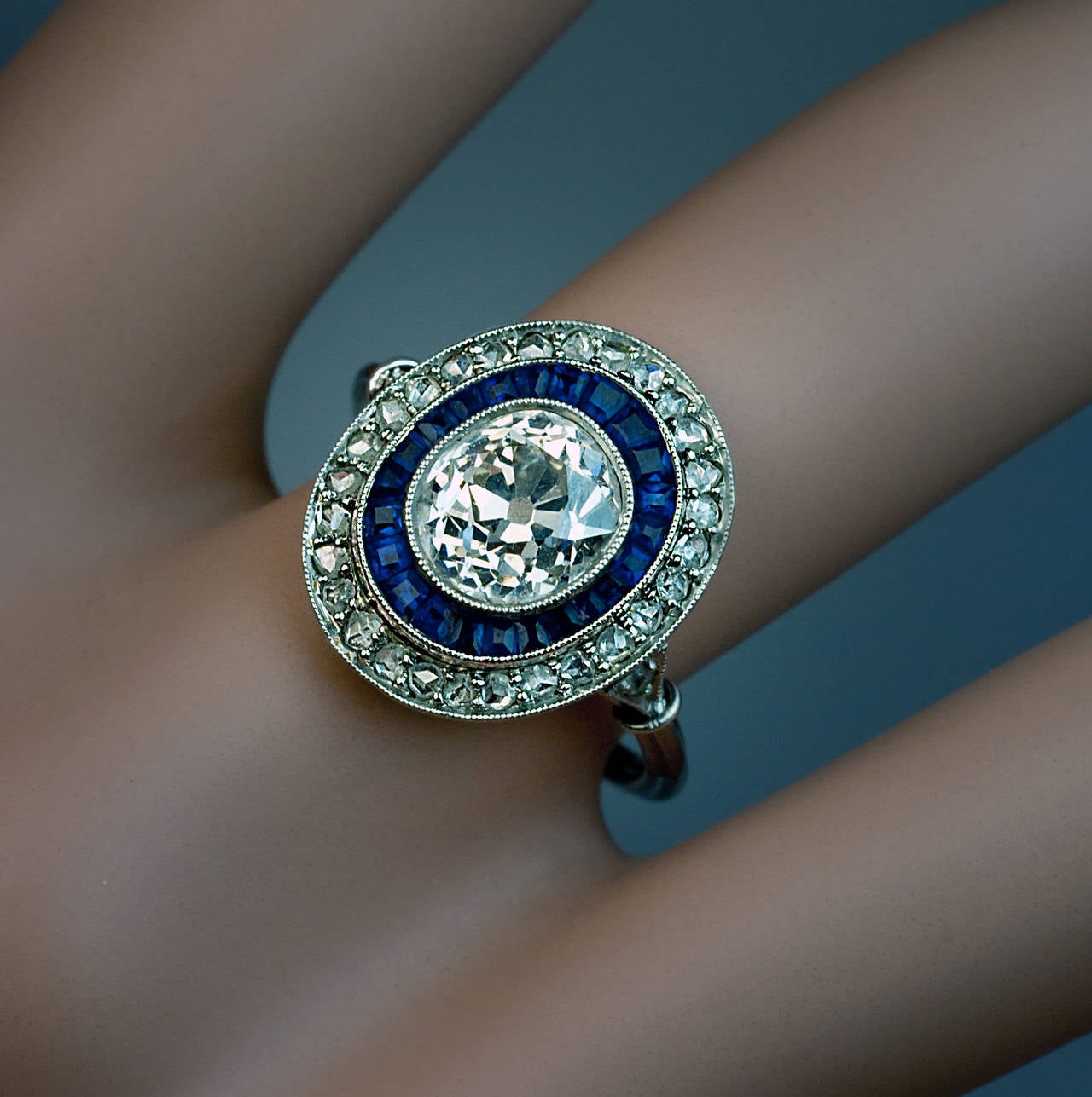 A platinum milgrain ring of an oval shape centered with a bezel set sparkling antique cushion cut diamond (7.3 x 6.8 x 4.85 mm, approximately 2 carats, K color, VS2 clarity) outlined by a row of tapered calibre cut deep blue sapphires and further