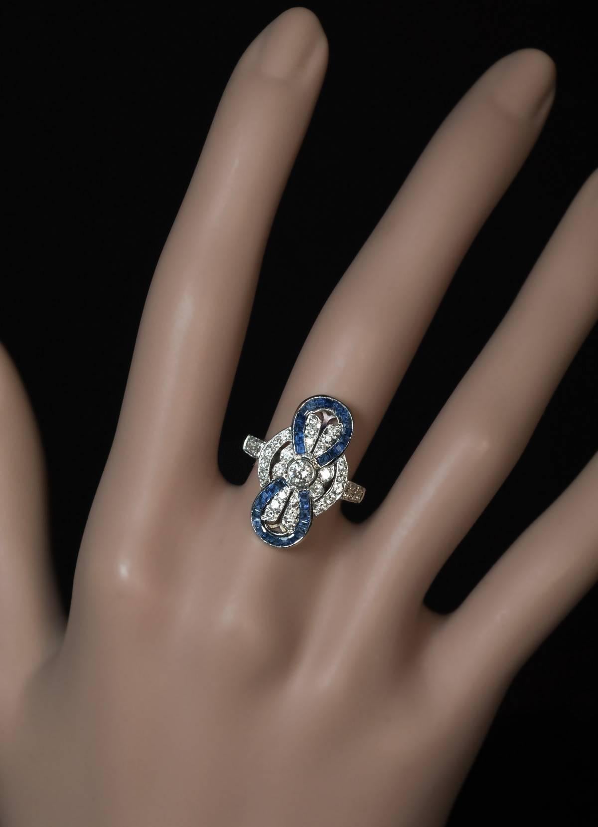 Circa 1950s

A white and yellow 18K gold bow motif ring is set with bright white brilliant cut diamonds and calibre cut (French cut) sapphires.

Estimated total diamond weight is 1 carat.

Size of the diamond and sapphire top 23 x 14 mm (15/16 x 1/2