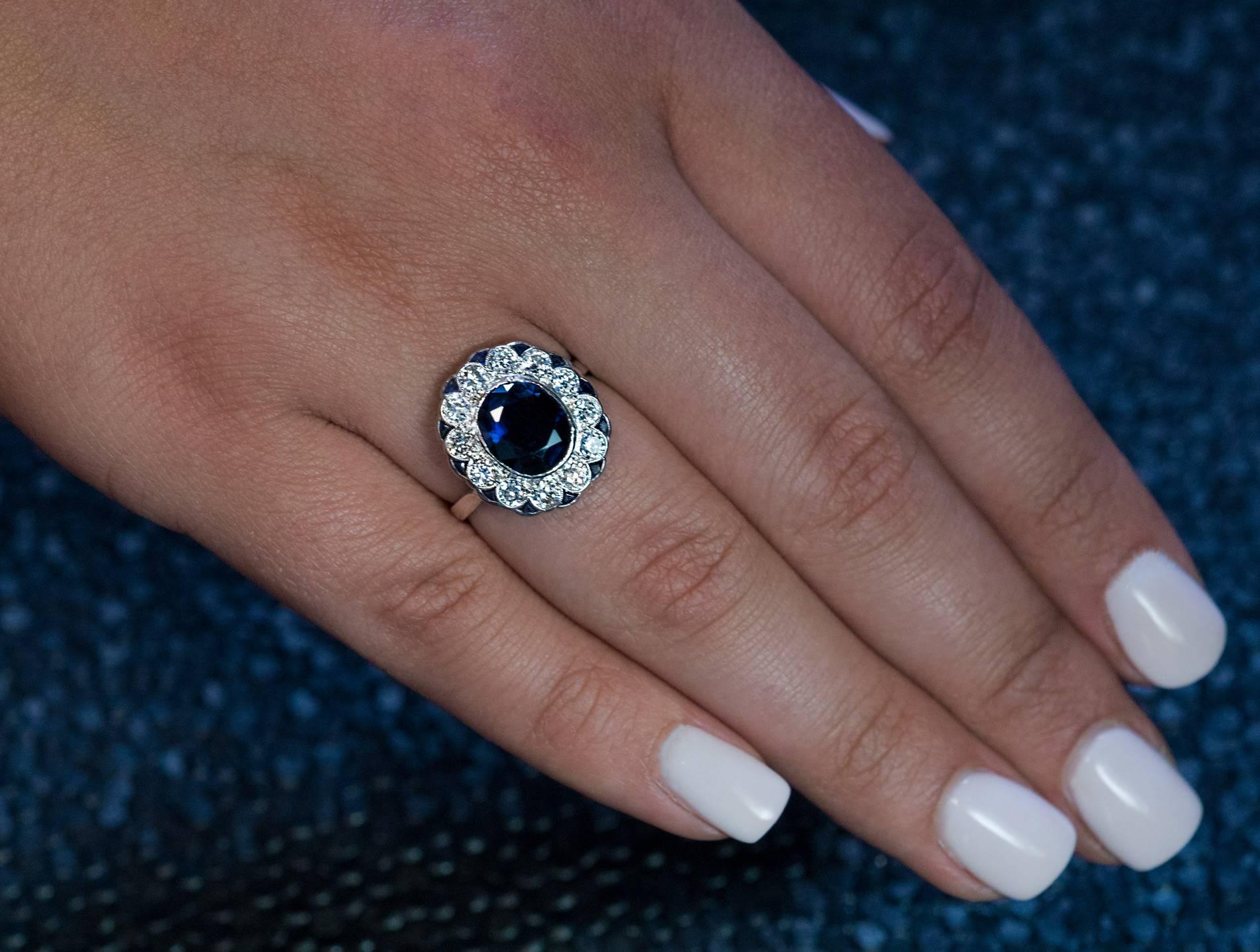 Circa 1950s
The 14K white gold ring is centered with a bezel set natural sapphire (9.2 x 7.8 x 4.6 mm, approximately 2.64 ct) surrounded by 12 bright white brilliant cut diamonds (F-G color, VS2-SI2 clarity), in turn, outlined by calibre cut